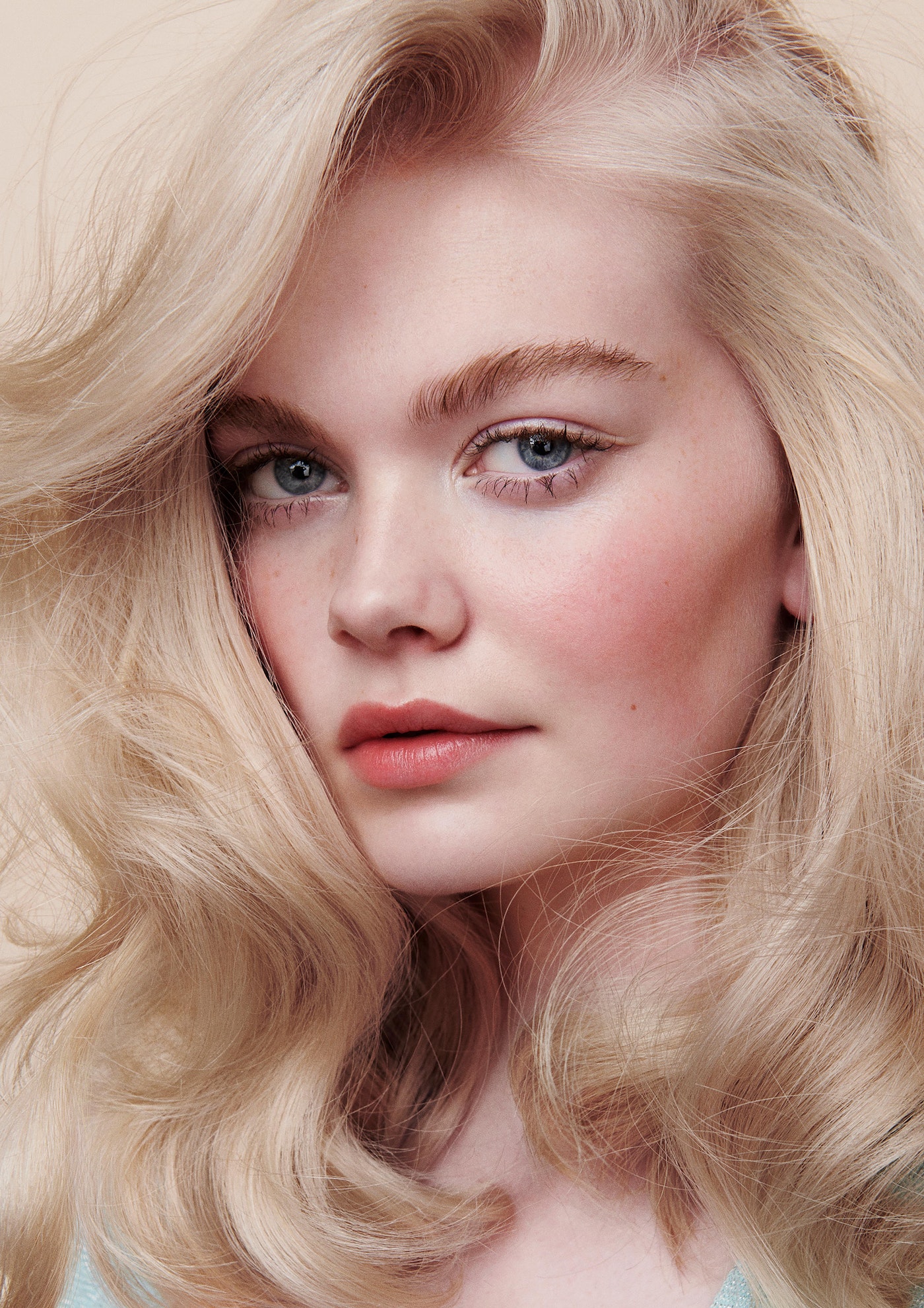 Close-up portrait of a young fair-skinned woman with wavy blonde hair and blue eyes in soft lighting against a neutral background.