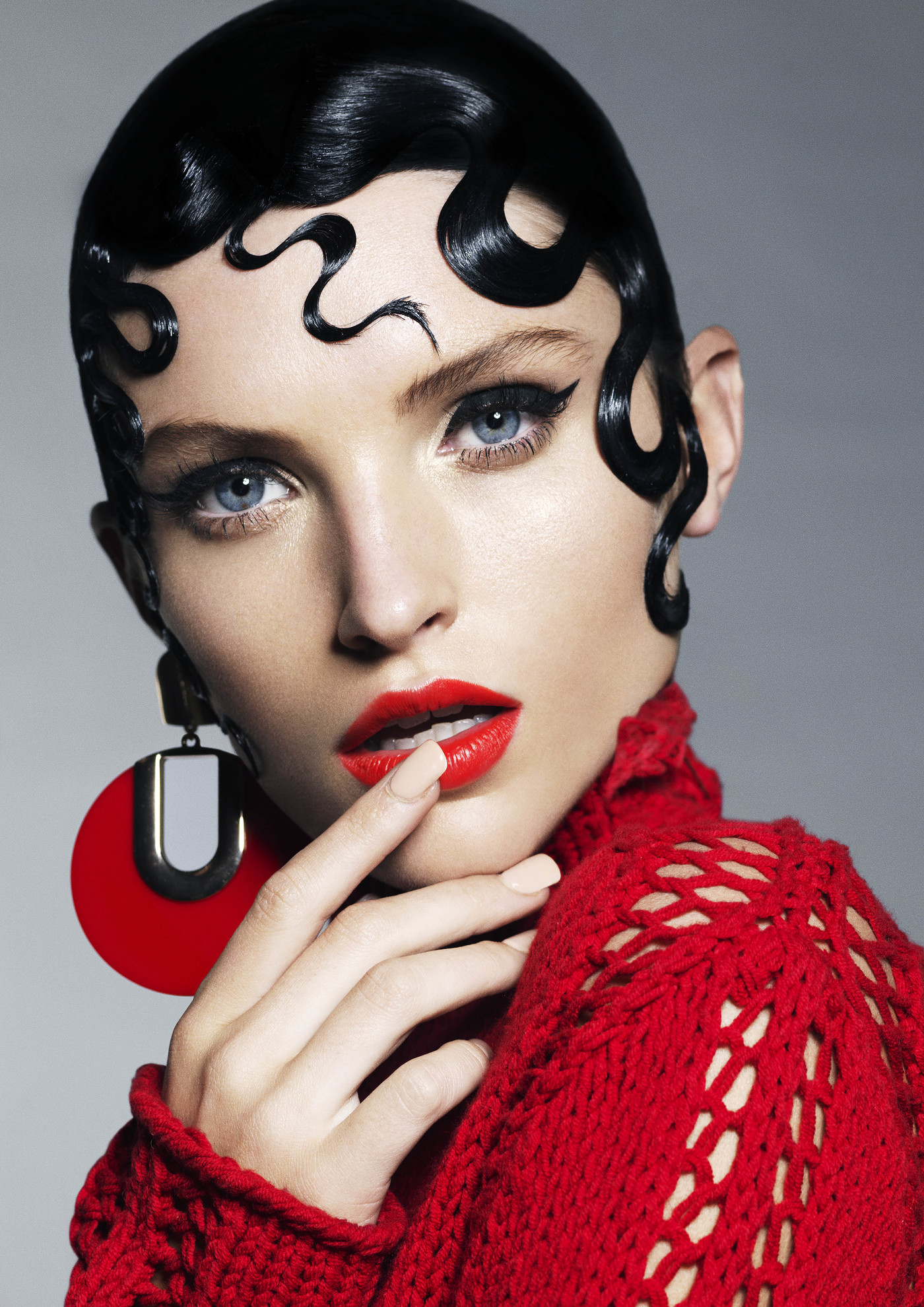 Close-up of a woman with dark wavy hair, red lipstick, and red knitwear. She is wearing large red earrings, posing in a studio.