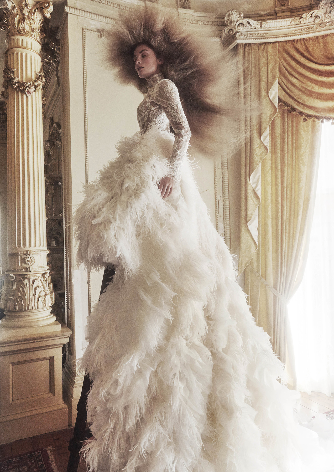 A woman in a white feathered gown stands in an ornate room with a high ceiling and columns, showcasing fashion and editorial style.