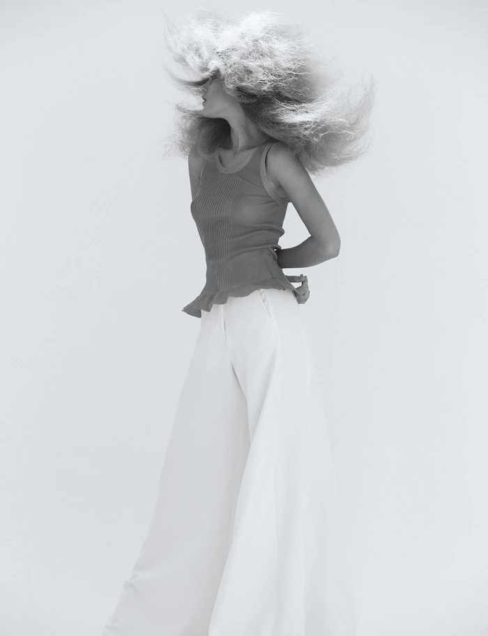 Monochrome portrait of a woman with voluminous textured hair wearing a sleeveless top and white trousers