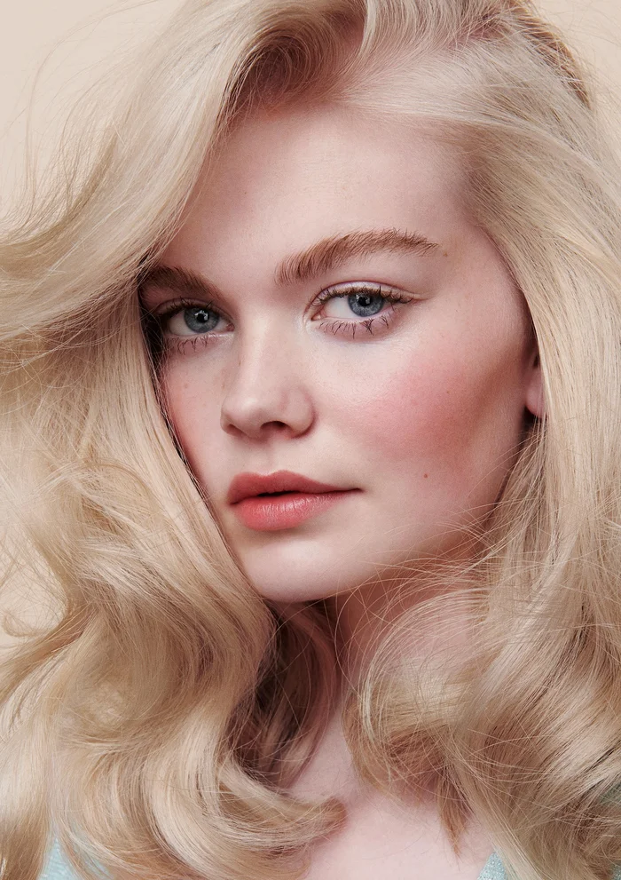 Close-up portrait of a blonde woman with voluminous wavy hair, soft neutral makeup, and blue eyes against a light background.