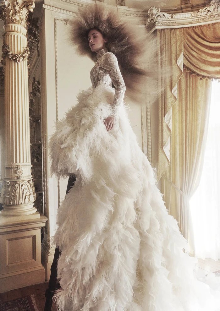 Model with voluminous feathered hair wearing an elaborate white lace and feather gown inside a grand room