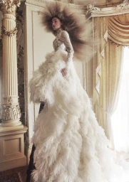 Model with voluminous feathered hair wearing an elaborate white lace and feather gown inside a grand room
