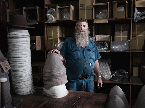 Mature bearded man poses near stacks of hats inside millinery workshop, surrounded by boxes and tools, creating a vintage feel.