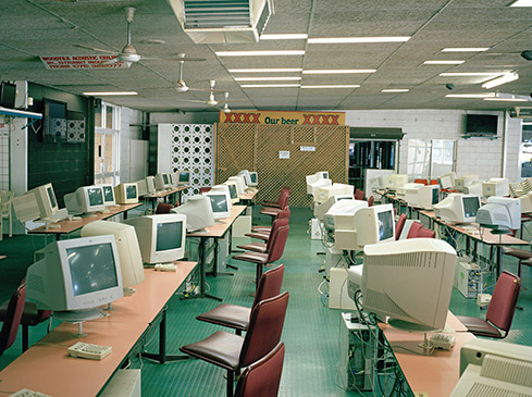 Rows of vintage computers, keyboards, and red chairs fill a brightly lit indoor room under a drop ceiling with XXXX beer signage.