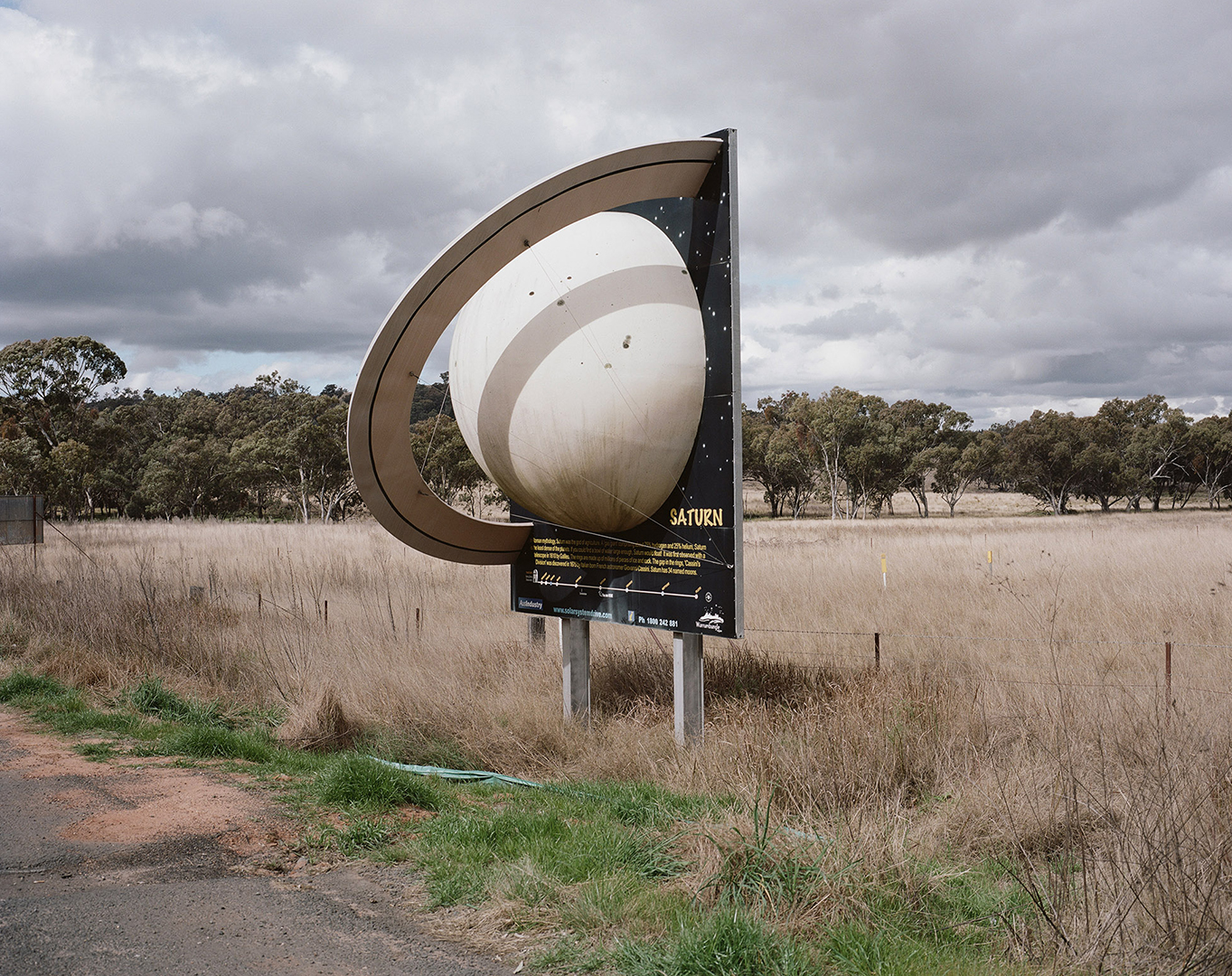 Outdoor photo of an artistic Saturn planet sign in a field under a cloudy sky, surrounded by dry grass and scattered trees.