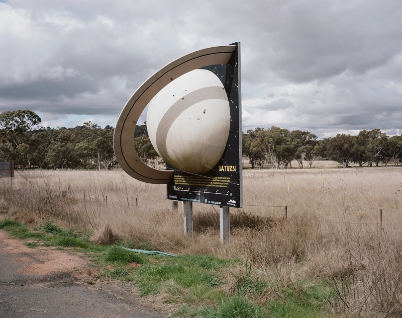 Outdoor photo of an artistic Saturn planet sign in a field under a cloudy sky, surrounded by dry grass and scattered trees.