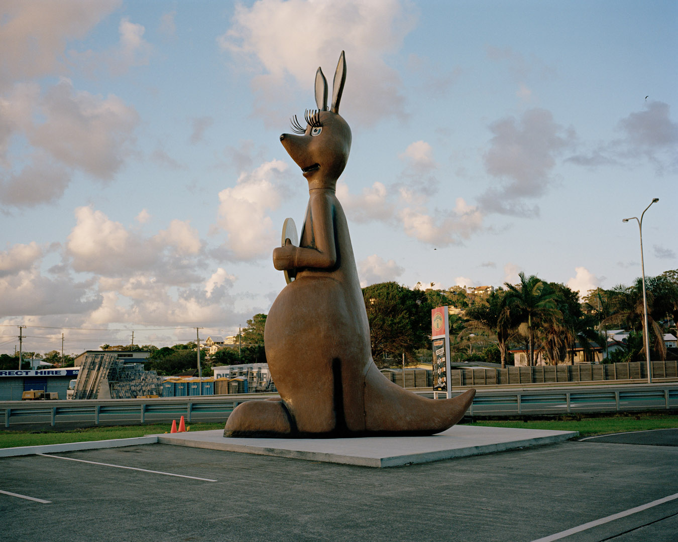 Large kangaroo statue in outdoor setting with cloudy skies; roadside attraction with Direct Hire business and palm trees in background.