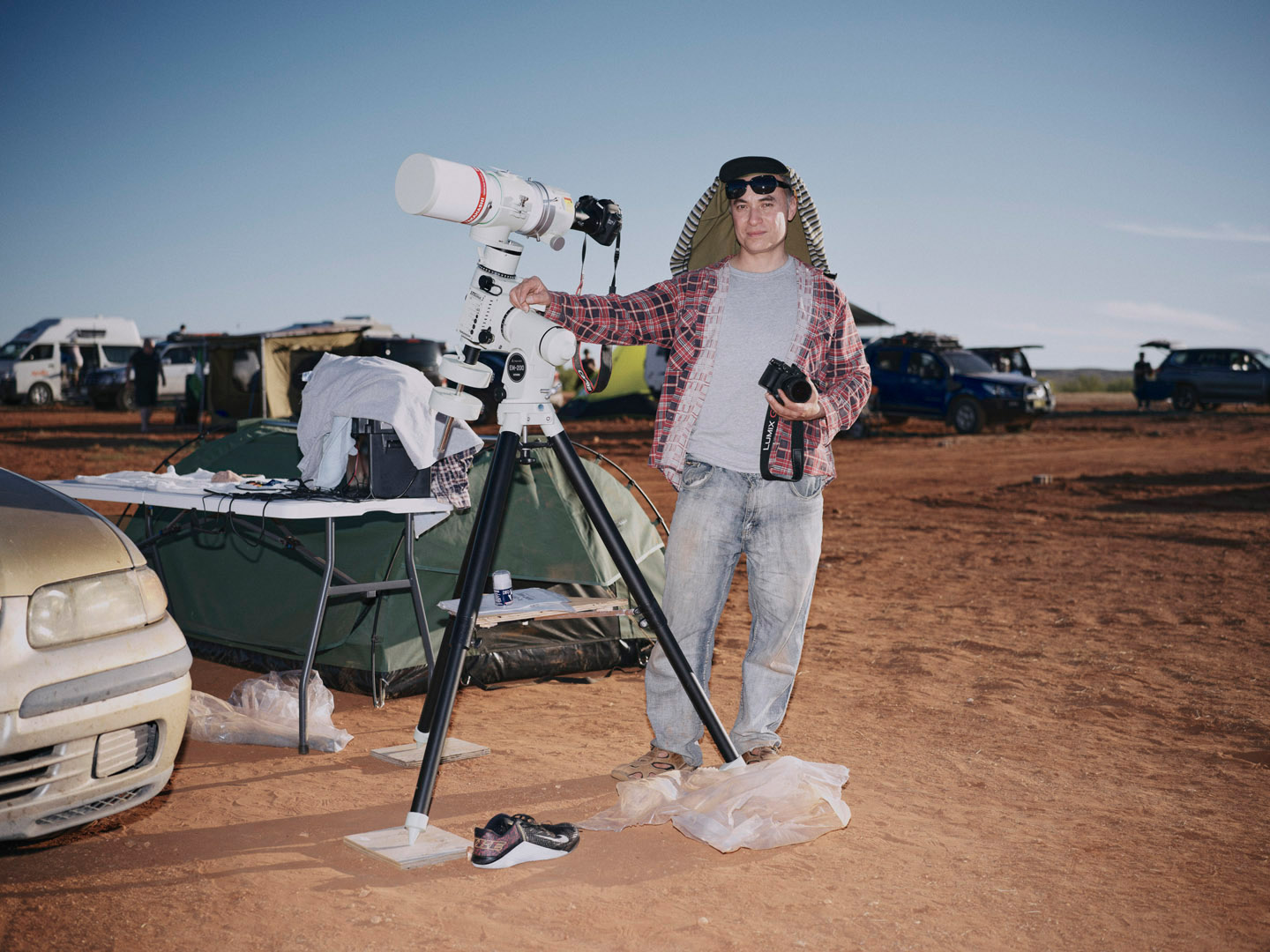 Man stands next to a telescope on a tripod at an outdoor campsite on a sunny day, with cars and tents in the background.