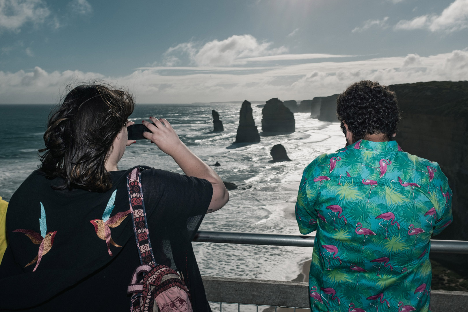 Two people admire coastal rock formations at Twelve Apostles Marine National Park in Victoria, Australia, under a partly cloudy sky.