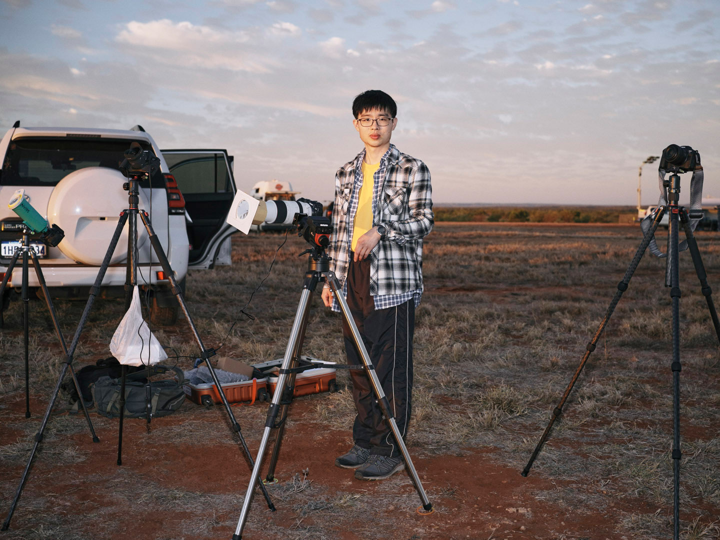 Man standing outdoors with cameras on tripods, cars, and open land at sunset. The photographer is an Asian man wearing glasses.
