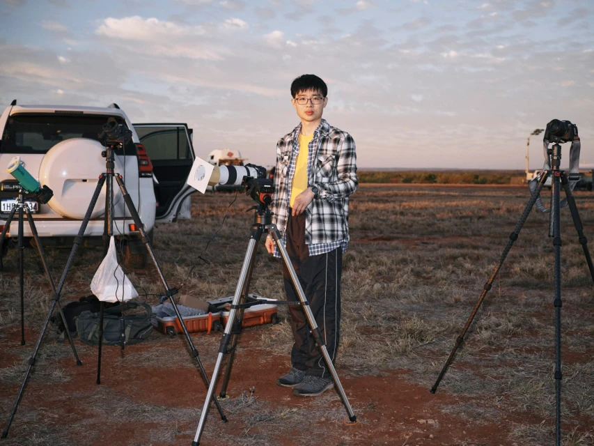 Man standing outdoors with cameras on tripods, cars, and open land at sunset. The photographer is an Asian man wearing glasses.