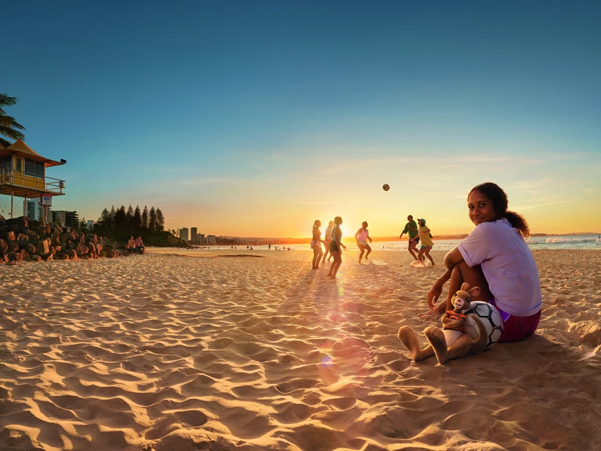 Teenage girl with a teddy bear poses on a sandy beach at sunset as a group plays soccer near a lifeguard tower.
