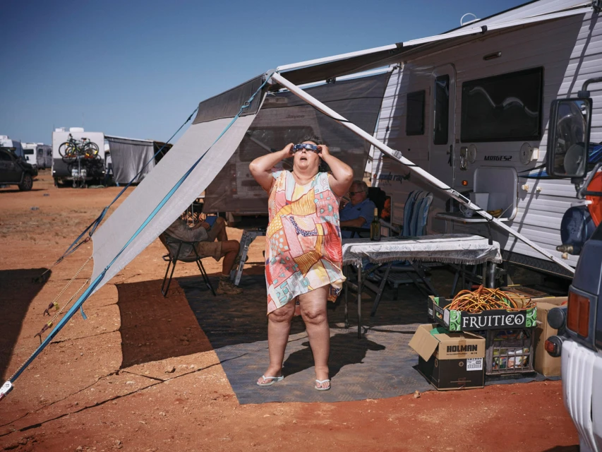 Woman in sunglasses stands outside RV under awning with boxes in a campground on a sunny day in a medium, eye-level shot.