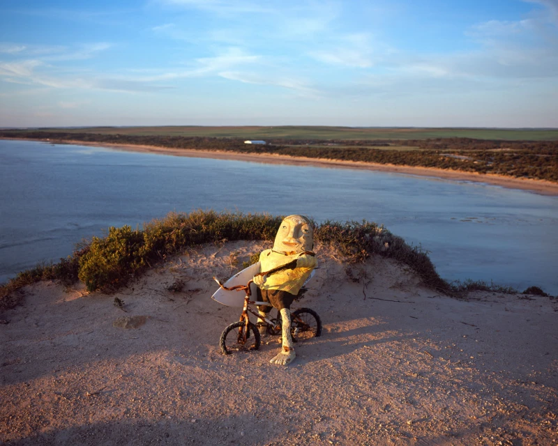 Quirky humanoid scarecrow figure rides a bicycle with a surfboard on a sandy dune overlooking ocean and coastal land, with a blue sky.
