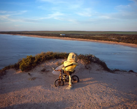 Quirky humanoid scarecrow figure rides a bicycle with a surfboard on a sandy dune overlooking ocean and coastal land, with a blue sky.