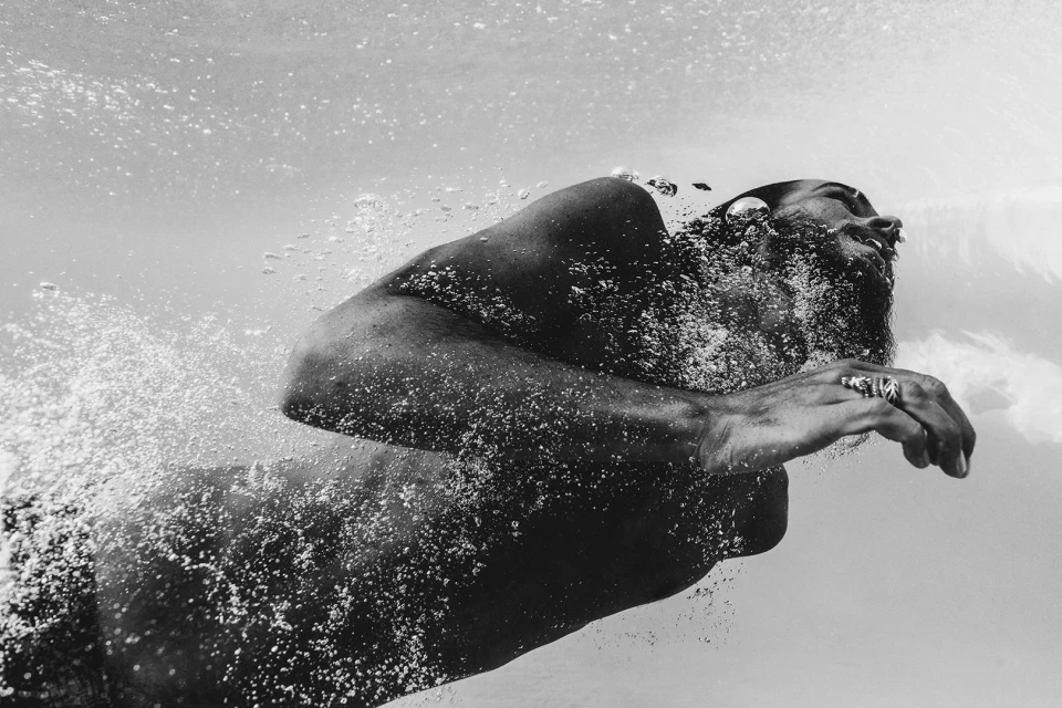 Monochrome portrait of an adult male underwater with a beard and rings, surrounded by bubbles in a clear body of water.