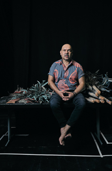 Portrait of a mature man with a bald head wearing a patterned shirt sitting in front of a pile of plant material and a black background.