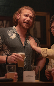 Two adult men at a bar with one man's hand on the other's shoulder, with drinks on the counter and a dark background, casual, friendly.