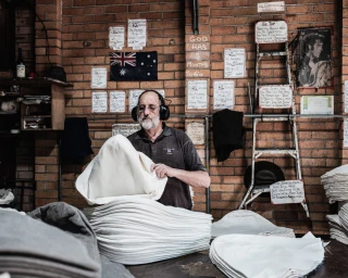 An older man wearing earmuffs sorting white fabric materials inside a brick workshop photographed by Trent Mitchell.