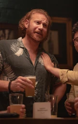 Red-haired man in a patterned short-sleeve shirt holding a beer glass in a bar, photographed by Trent Mitchell.
