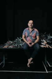 Bald man seated barefoot on a black table with plants, wearing a patterned blue shirt and dark trousers against a dark background.