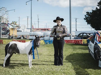 Woman in cowboy hat and patterned shirt standing on grass holding lead of a small black and white pony