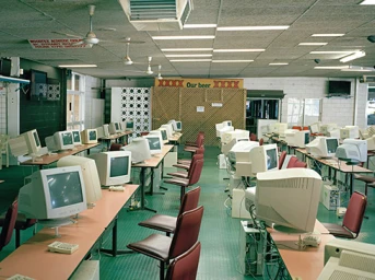 Rows of vintage desktop computers and CRT monitors on tables in an expansive office space with a green floor and acoustic ceiling panels.