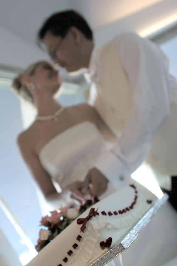 Newlywed couple cut wedding cake together at indoor venue with natural light. Bride in gown and groom in tuxedo celebrate special day.