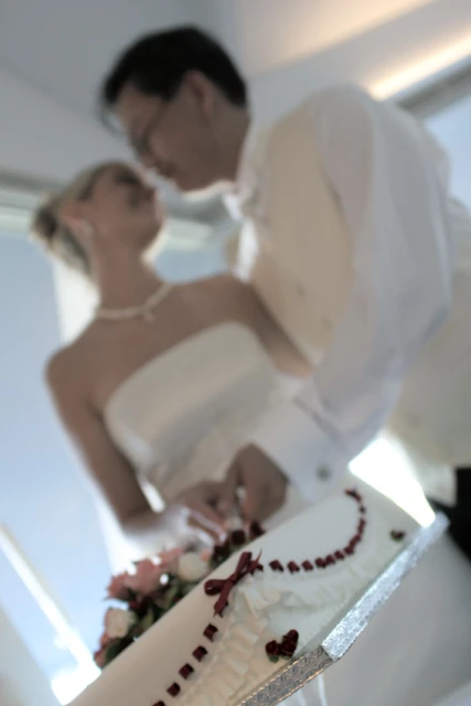 Newlywed couple cut wedding cake together at indoor venue with natural light. Bride in gown and groom in tuxedo celebrate special day.