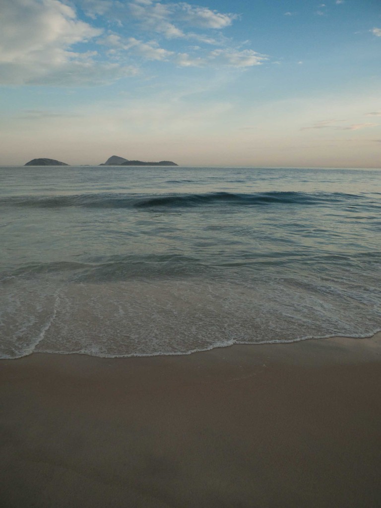 Scenic view of a tranquil beach at sunset with gentle waves meeting the sandy shore under a partly cloudy sky and distant islands.