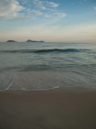 Scenic view of a tranquil beach at sunset with gentle waves meeting the sandy shore under a partly cloudy sky and distant islands.