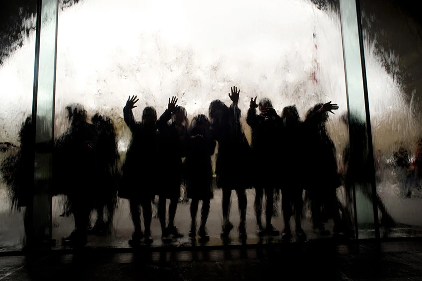 A group of silhouetted people, including children, gather behind a glass water feature, creating a dynamic and blurred visual effect.