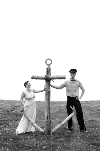 Monochrome photo of a couple posing with a wooden anchor on a grassy field under a bright sky, creating a vintage, nautical-themed portrait.