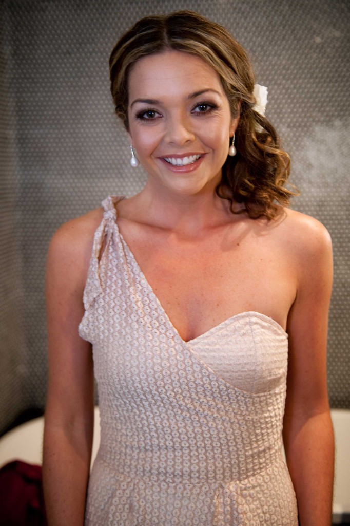 Portrait of a smiling woman with brown hair and a cream-colored one-shoulder dress, wearing pearl earrings and a white flower hair accessory.