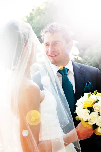 A bride and groom share a loving look in an outdoor wedding scene, the bride in a veil, the groom in a suit with a teal tie, and white and yellow roses.