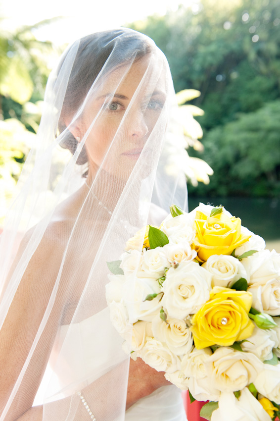 Woman in a white wedding dress with a veil poses outdoors holding a bouquet of white and yellow flowers, with a forest in the background.