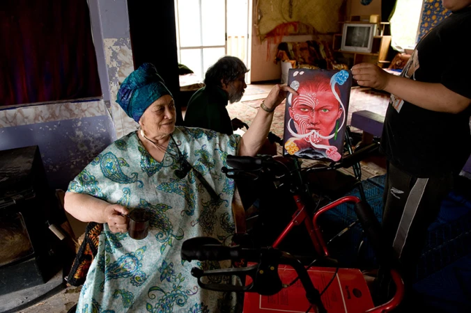 Elderly woman in patterned dress sits with a cup, holding up a colorful portrait, other people are visible in the background indoors.