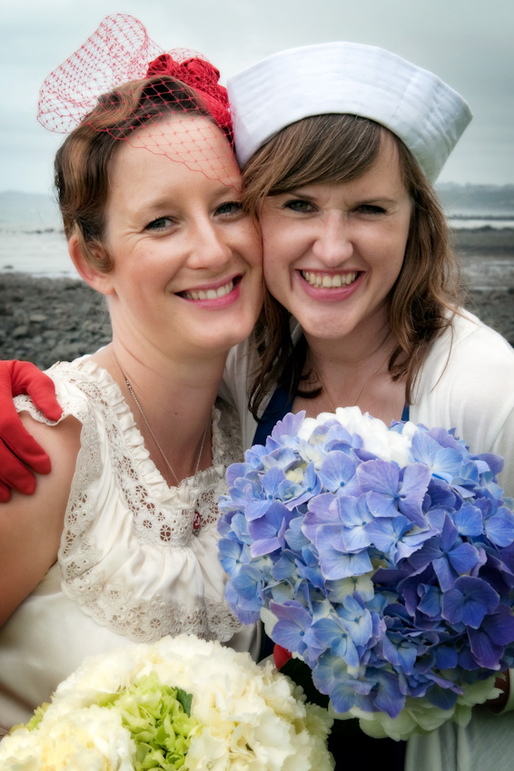 Two women in vintage attire pose with hydrangea bouquets on a cloudy beach. One wears a red hat with netting, the other a white sailor hat.