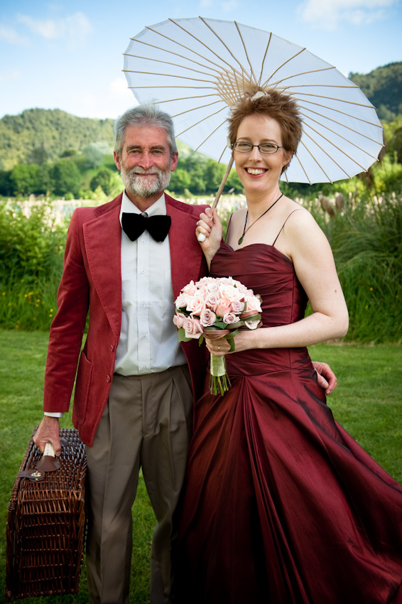 Man and woman in formal attire pose outdoors with parasol and picnic basket, smiling against a backdrop of greenery and rolling hills.