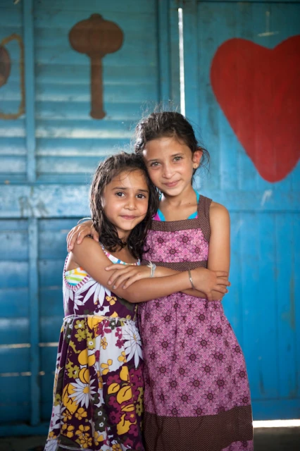 Two young girls with arms around each other stand in front of a blue wall with folk art. They wear summer dresses and smile.