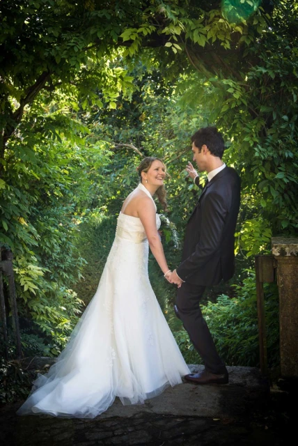 A smiling bride in a white dress and a groom in a black suit hold hands in a lush garden setting surrounded by vibrant green foliage.