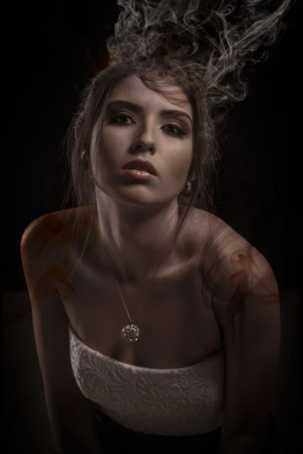 Portrait of a young woman with smoke details against a dark background. She's wearing jewelry and a white dress, with soft, moody lighting.