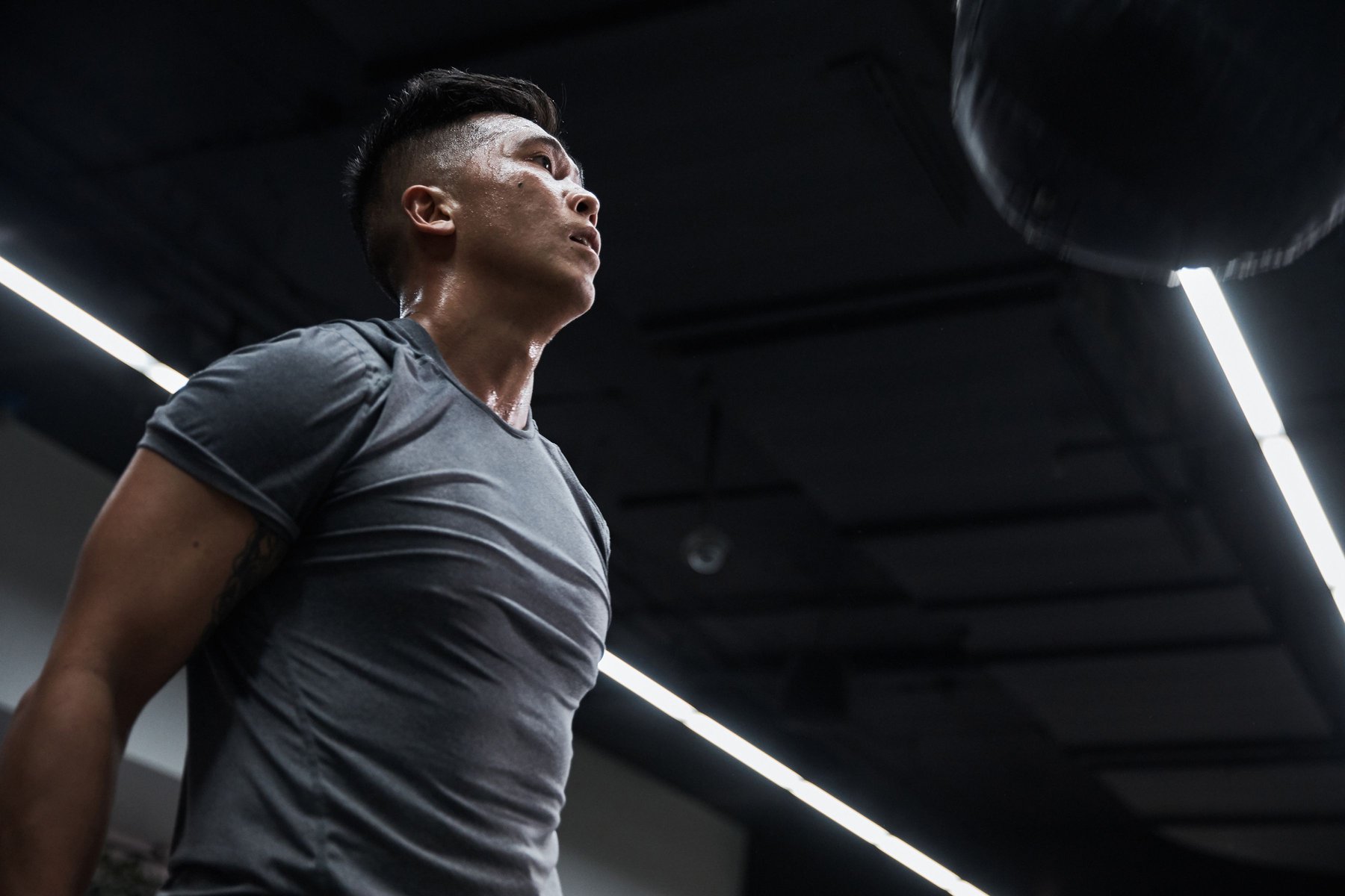 Sweaty muscular man in gray workout shirt looks up at speed bag hanging in dark gym with bright ceiling lights during training session.