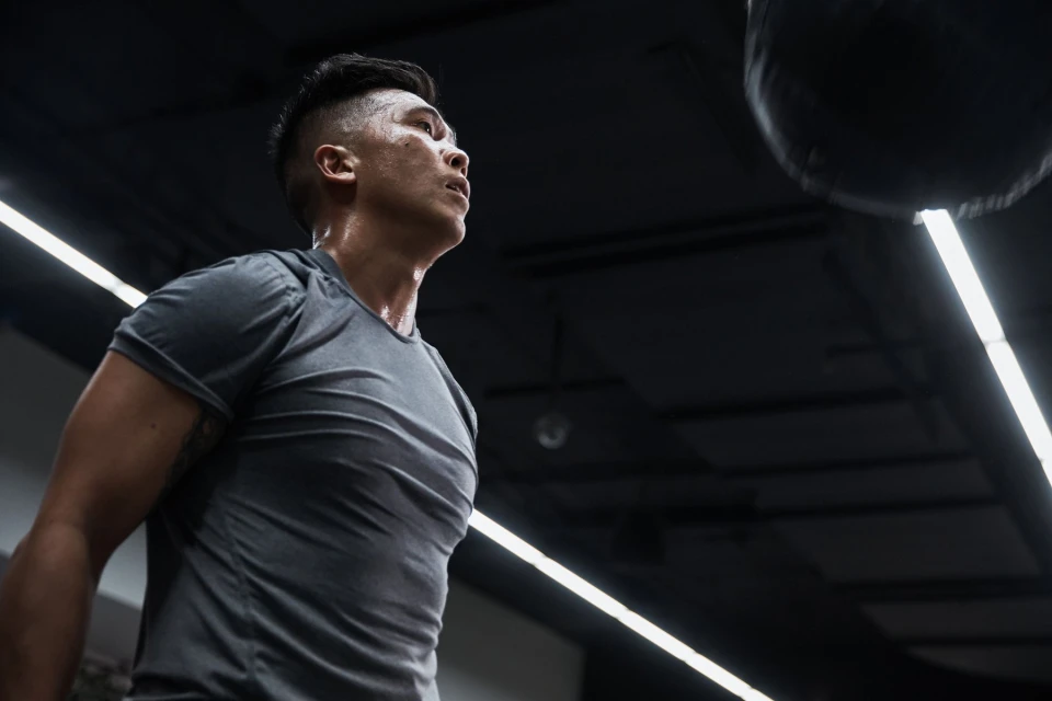 Sweaty muscular man in gray workout shirt looks up at speed bag hanging in dark gym with bright ceiling lights during training session.