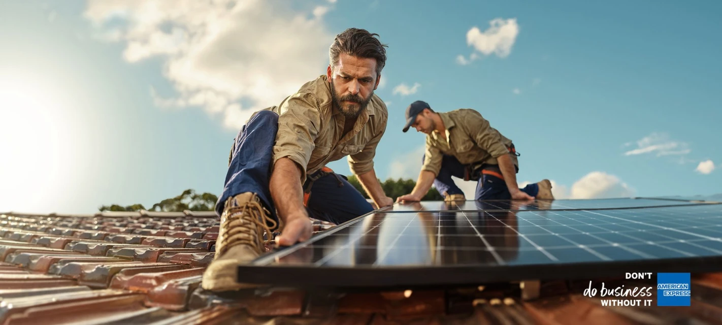 Two men installing solar panels on a rooftop under a partly cloudy sky, wearing work clothes and using equipment for the installation.