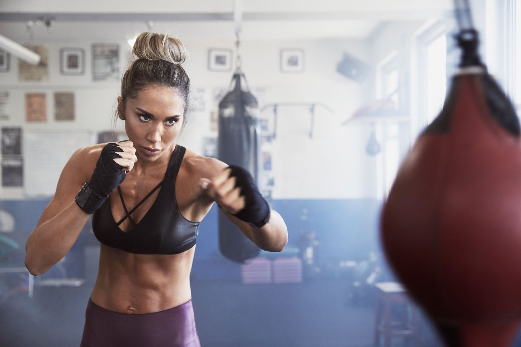 A muscular woman with blonde hair in a bun is throwing a punch while wearing boxing wraps in a gymnasium setting.