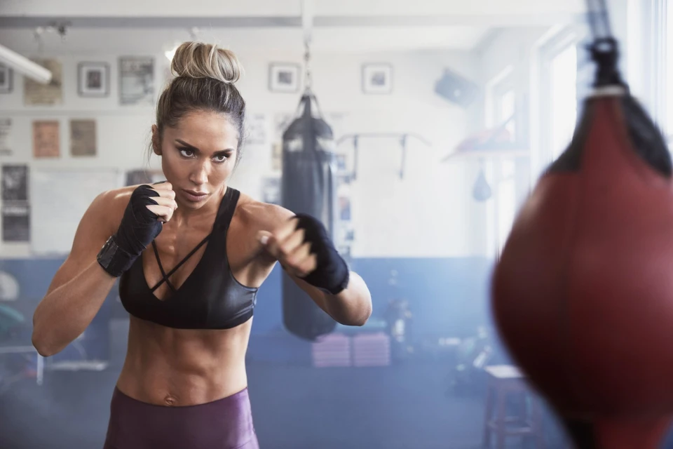 A muscular woman with blonde hair in a bun is throwing a punch while wearing boxing wraps in a gymnasium setting.