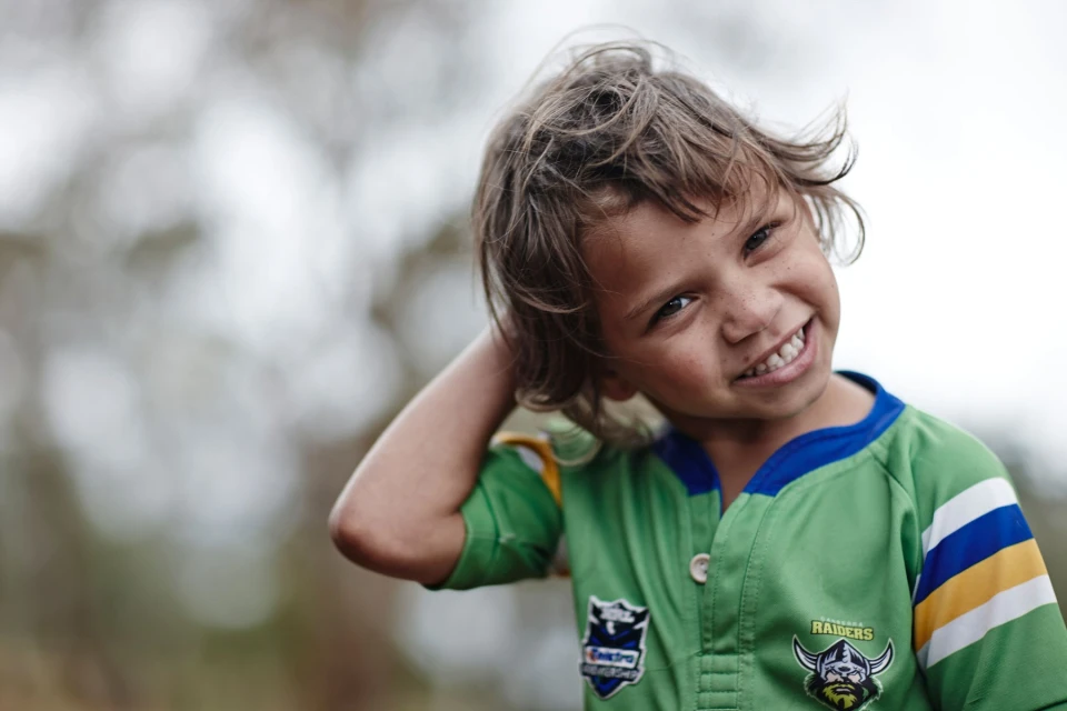Close-up of a smiling child with tousled brown hair, wearing a green and blue sports jersey, blurred background.