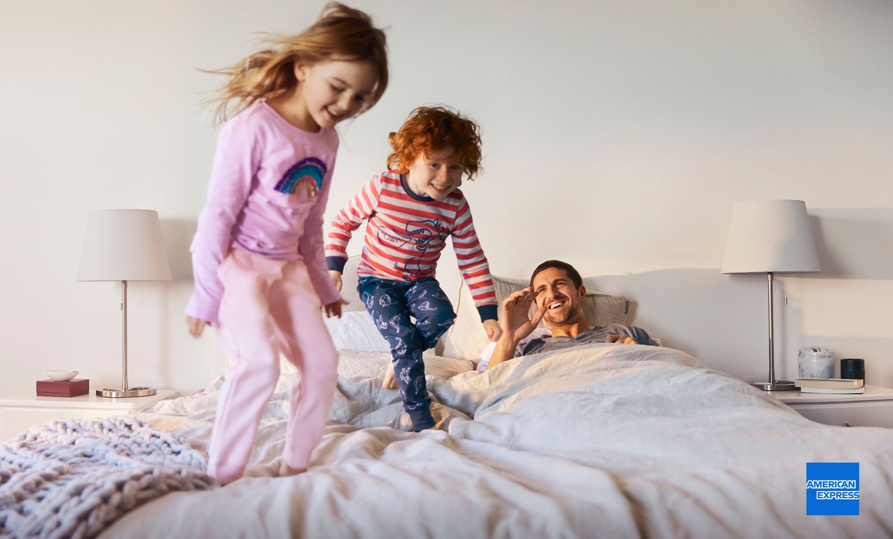 Two children jumping on a bed with their dad smiling in the background, illuminated by lamps on the nightstands in a home setting.